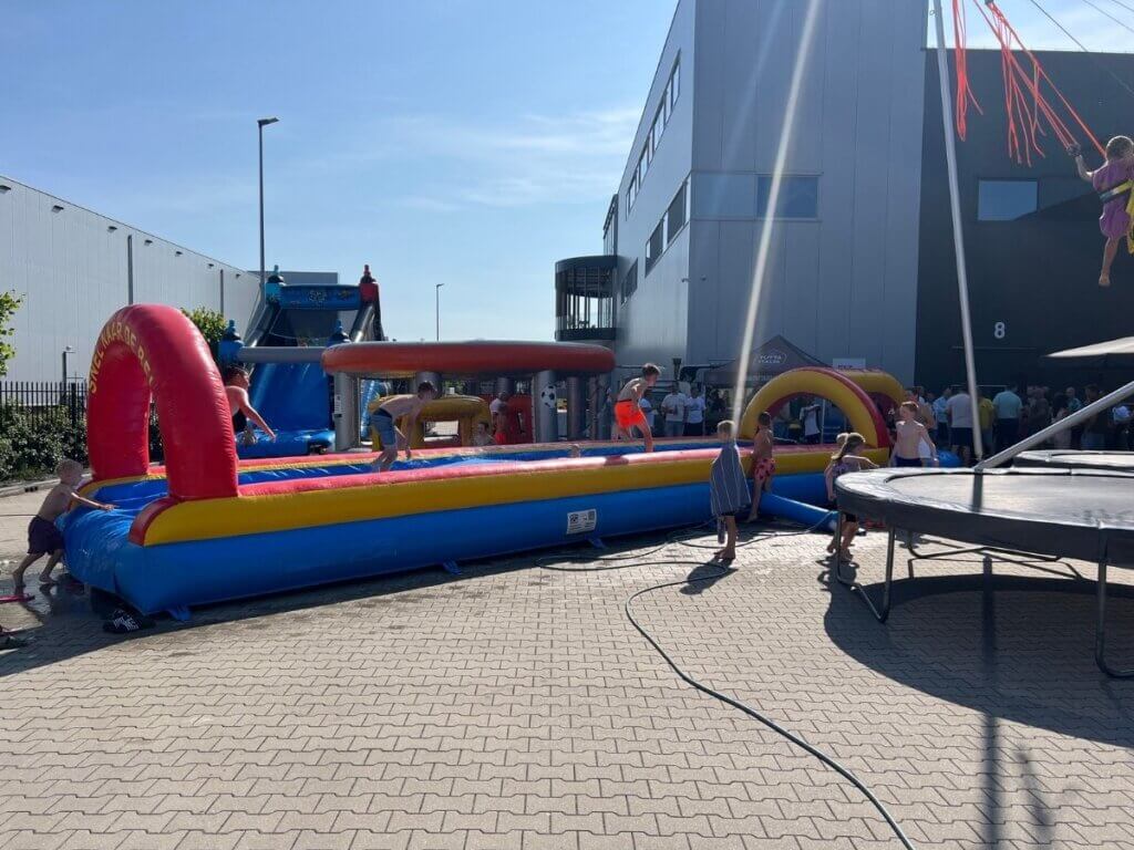 Kinderen spelen op een buikschuifbaan en stormbaan tijdens een zomerfeest bij een bedrijf, terwijl volwassenen op de achtergrond genieten van een drankje.