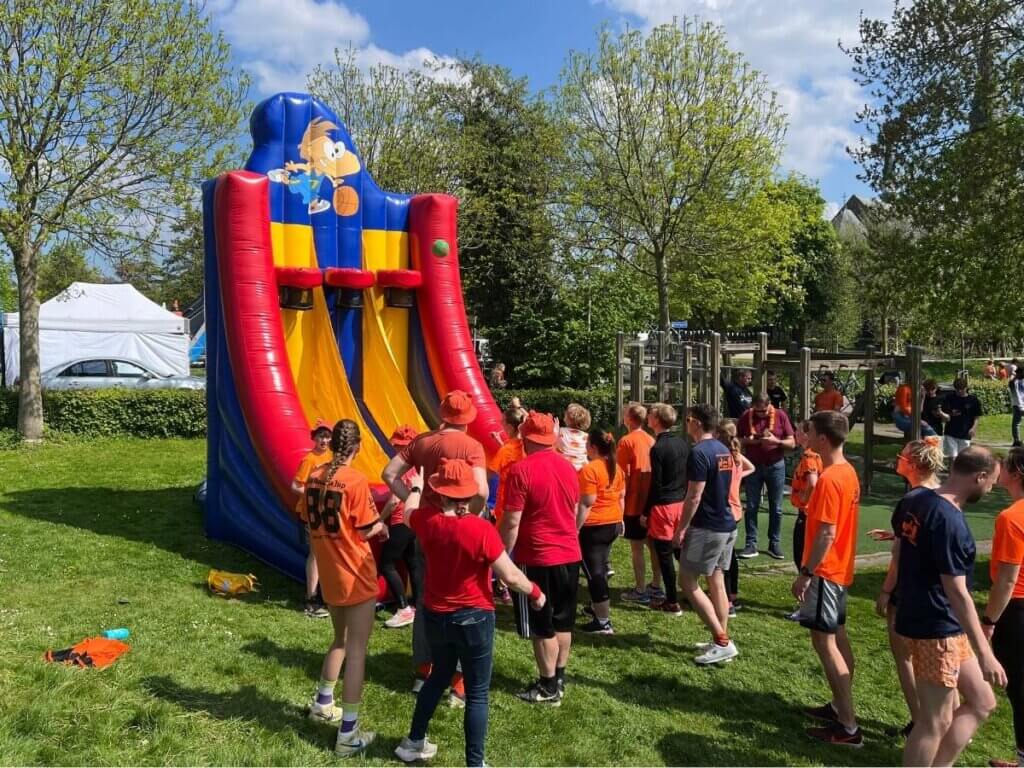 Deelnemers in oranje kleding wachten in de rij voor een groot springkussen basketbalspel tijdens een Koningsdag evenement.