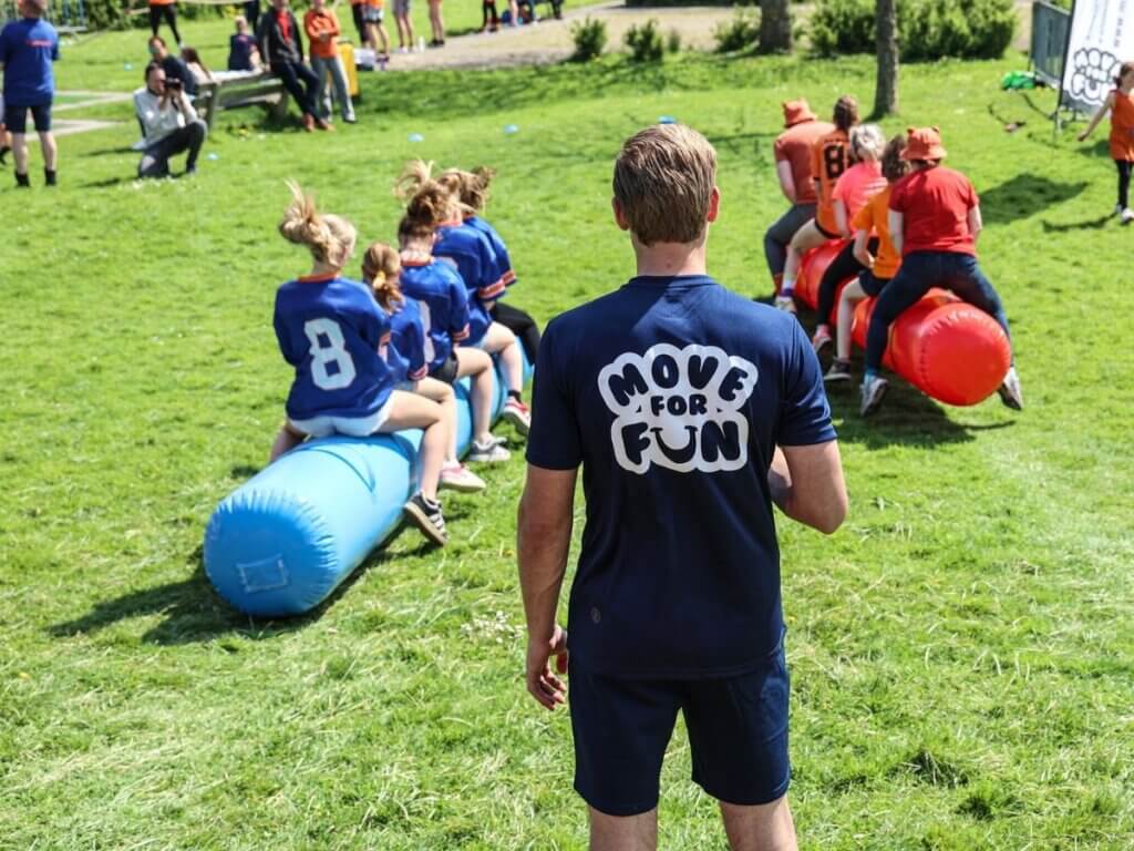 Begeleider van Move For Fun kijkt toe terwijl twee teams deelnemen aan een teamrace op opblaasbare rollen tijdens een Koningsdag evenement.